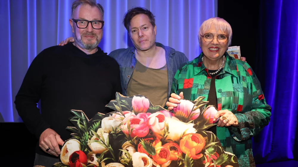 Über ein festliches Blumenbouguet aus Pappe hinweg schauen Holger Bergmann, Milo Rau und Claudia Roth in die Kamera.