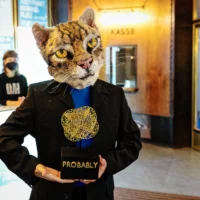 A performer in a black suit and a cat's head holds a trophy in front of him in his hands. The words Probably can be read in the base. In the background you can see the foyer of HAU 1 with an employee of the house.