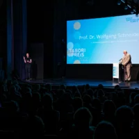 Prof. Dr. Wolfgang Schneider gives his opening speech on the stage of HAU1 in front of a big screen. On the left edge of the picture you can see the DGS translator of the evening.